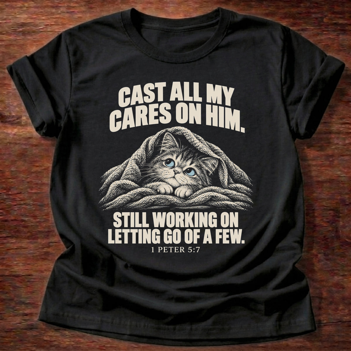 Letting Go Again T-Shirt in Black featuring a sleepy cat tucked under a blanket and 1 Peter 5:7 surrender humor design