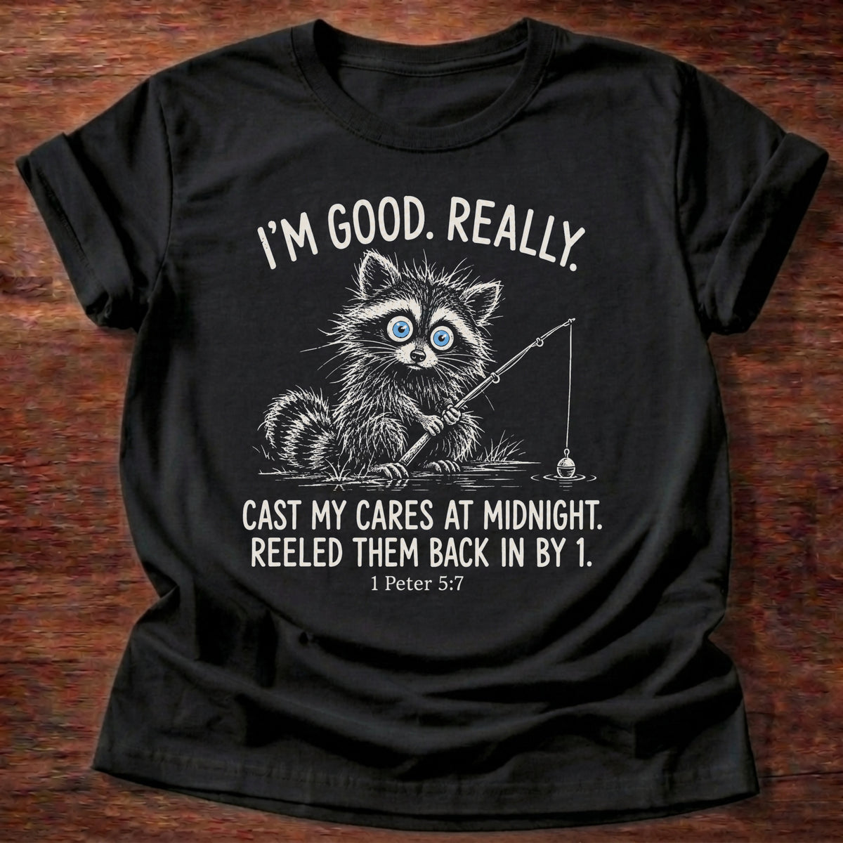 Black t-shirt printed with a raccoon holding a fishing rod graphic and “I'm good. Really. Cast my cares at midnight. Reeled them back in by 1.” 1 Peter 5:7 Christian shirt