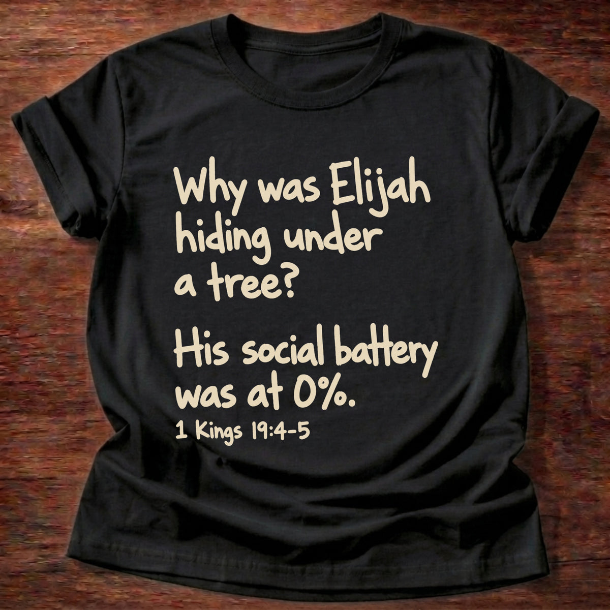 Black t-shirt printed with a simple hand-lettered Bible joke graphic and “Why was Elijah hiding under a tree? His social battery was at 0%.” 1 Kings 19:4-5 Christian shirt