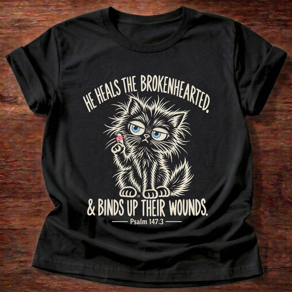 Black t-shirt printed with a scruffy fluffy white kitten holding a tiny pink heart graphic and “He heals the brokenhearted & binds up their wounds” Psalm 147:3 faith based gift