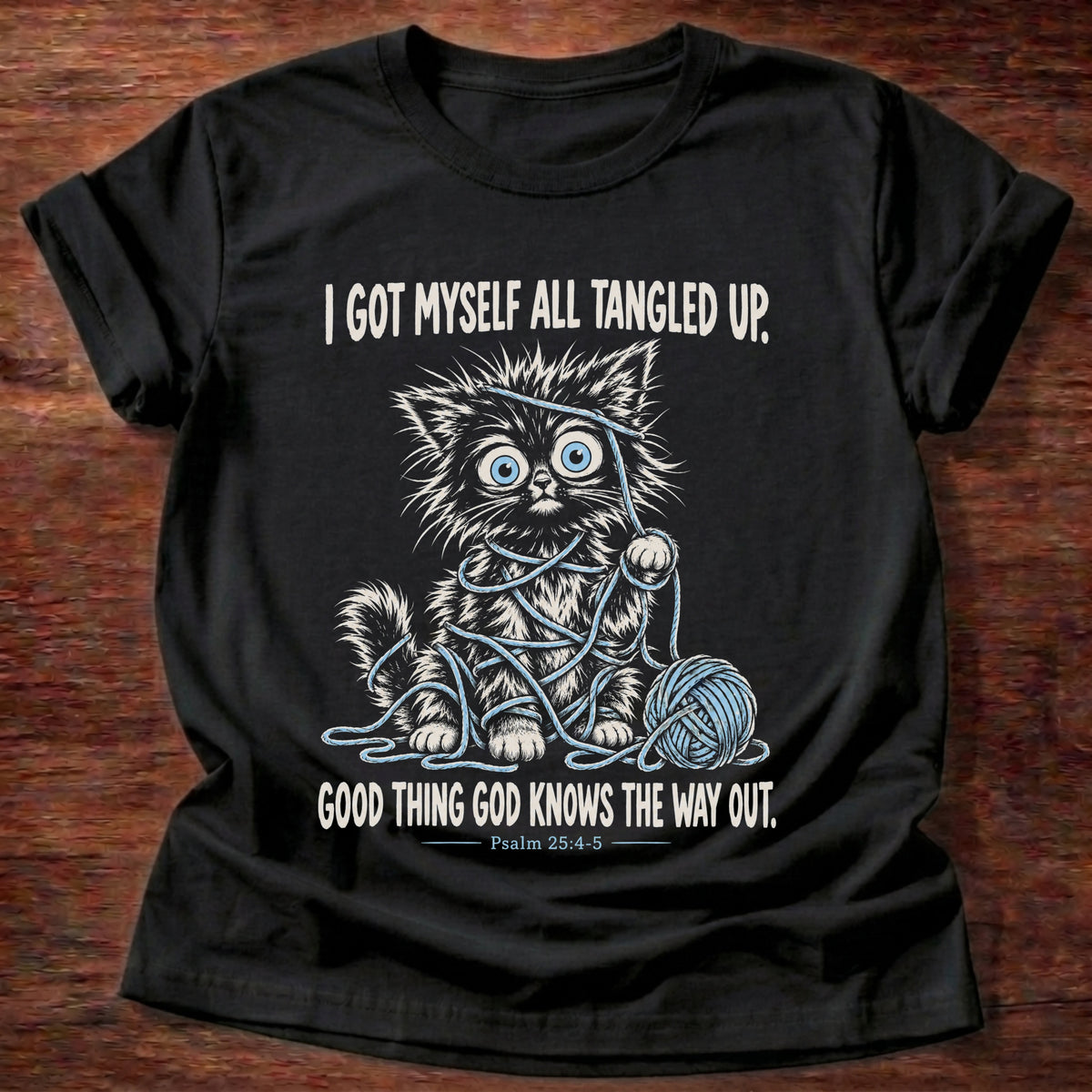 Black t-shirt printed with a wide-eyed kitten tangled up in blue yarn next to a yarn ball graphic and “I got myself all tangled up. Good thing God knows the way out.” Psalm 25:4-5 faith based gift
