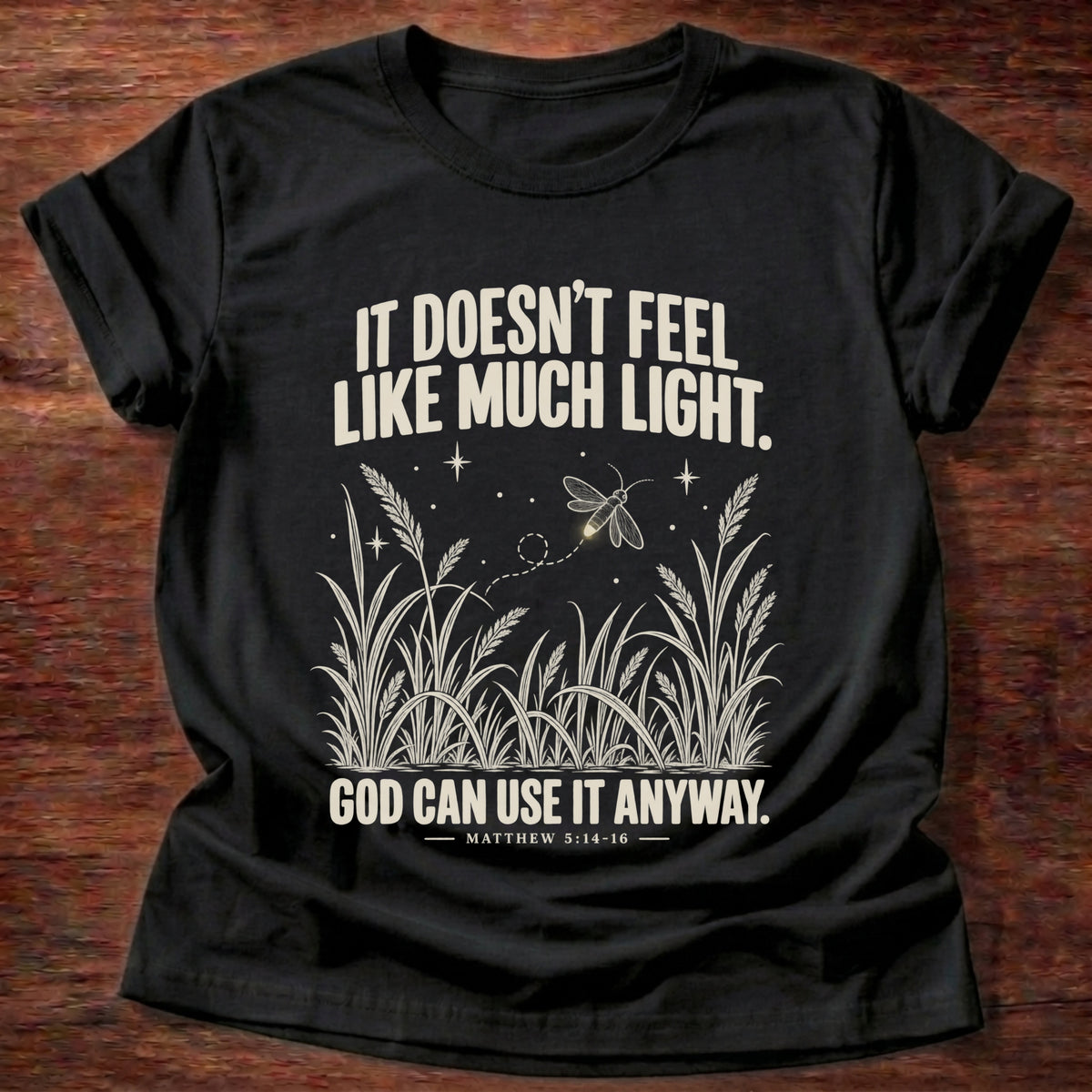 Black t-shirt printed with a firefly glowing in a dark field of grass graphic and 'It doesn't feel like much light. God can use it anyway.' Matthew 5:14-16 faith based gift