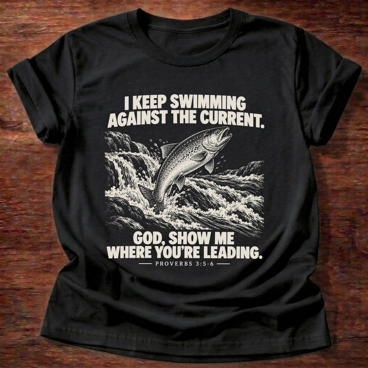 Black t-shirt printed with a fish leaping against a rushing current graphic and 'I keep swimming against the current. God, show me where you're leading.' Proverbs 3:5-6 faith based gift