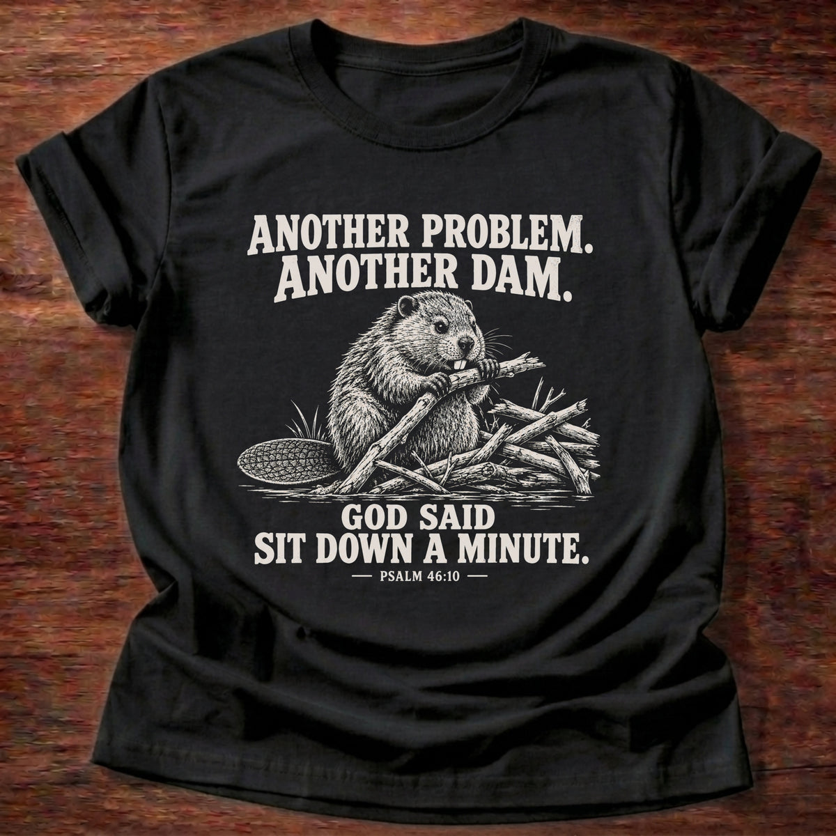 Black t-shirt printed with a beaver mid-build with another branch graphic and 'Another problem. Another dam. God said sit down a minute.' Psalm 46:10 faith based gift