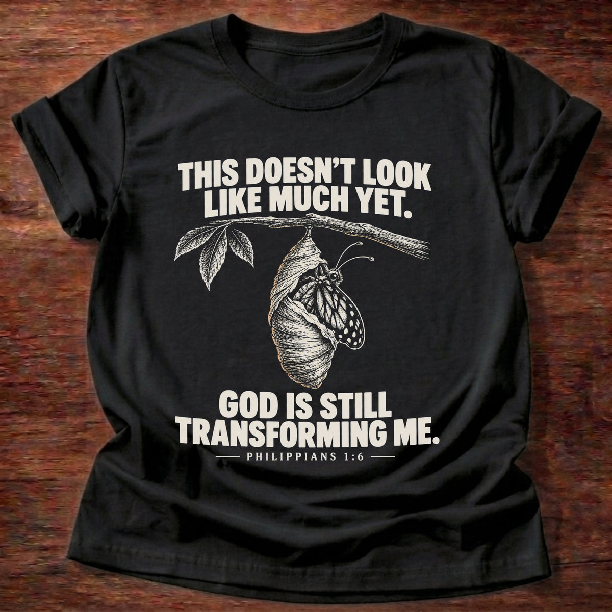 Black t-shirt printed with a chrysalis hanging from a branch graphic and 'This doesn't look like much yet. God is still transforming me.' Philippians 1:6 faith based gift