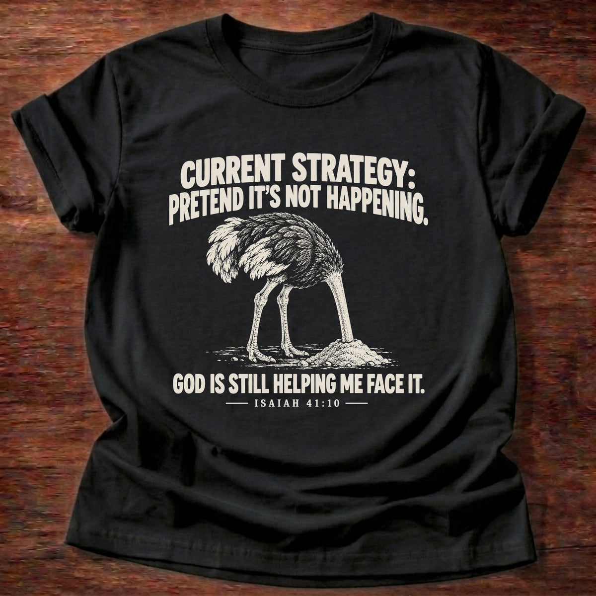 Black t-shirt printed with an ostrich with its head buried in the sand graphic and 'Current strategy: pretend it's not happening. God is still helping me face it.' Isaiah 41:10 faith based gift