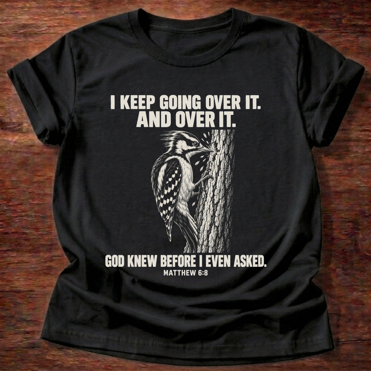 Black t-shirt printed with a woodpecker drumming the same spot on a tree graphic and 'I keep going over it. And over it. God knew before I even asked.' Matthew 6:8 faith based gift
