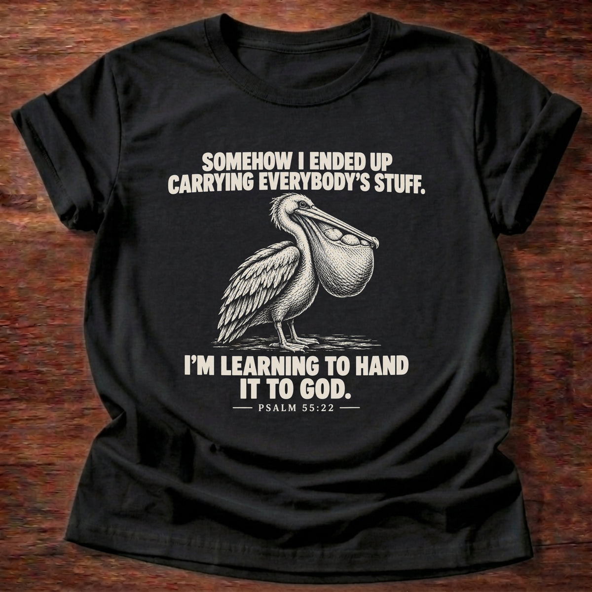 Black t-shirt printed with a pelican with a pouch full of fish graphic and 'Somehow I ended up carrying everybody's stuff. I'm learning to hand it to God.' Psalm 55:22 faith based gift