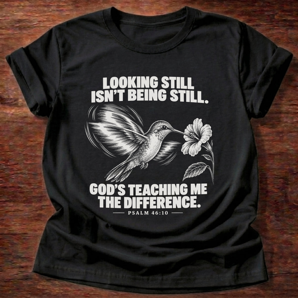 Black t-shirt printed with a hummingbird hovering beside a flower graphic and 'Looking still isn't being still. God's teaching me the difference.' Psalm 46:10 faith based gift