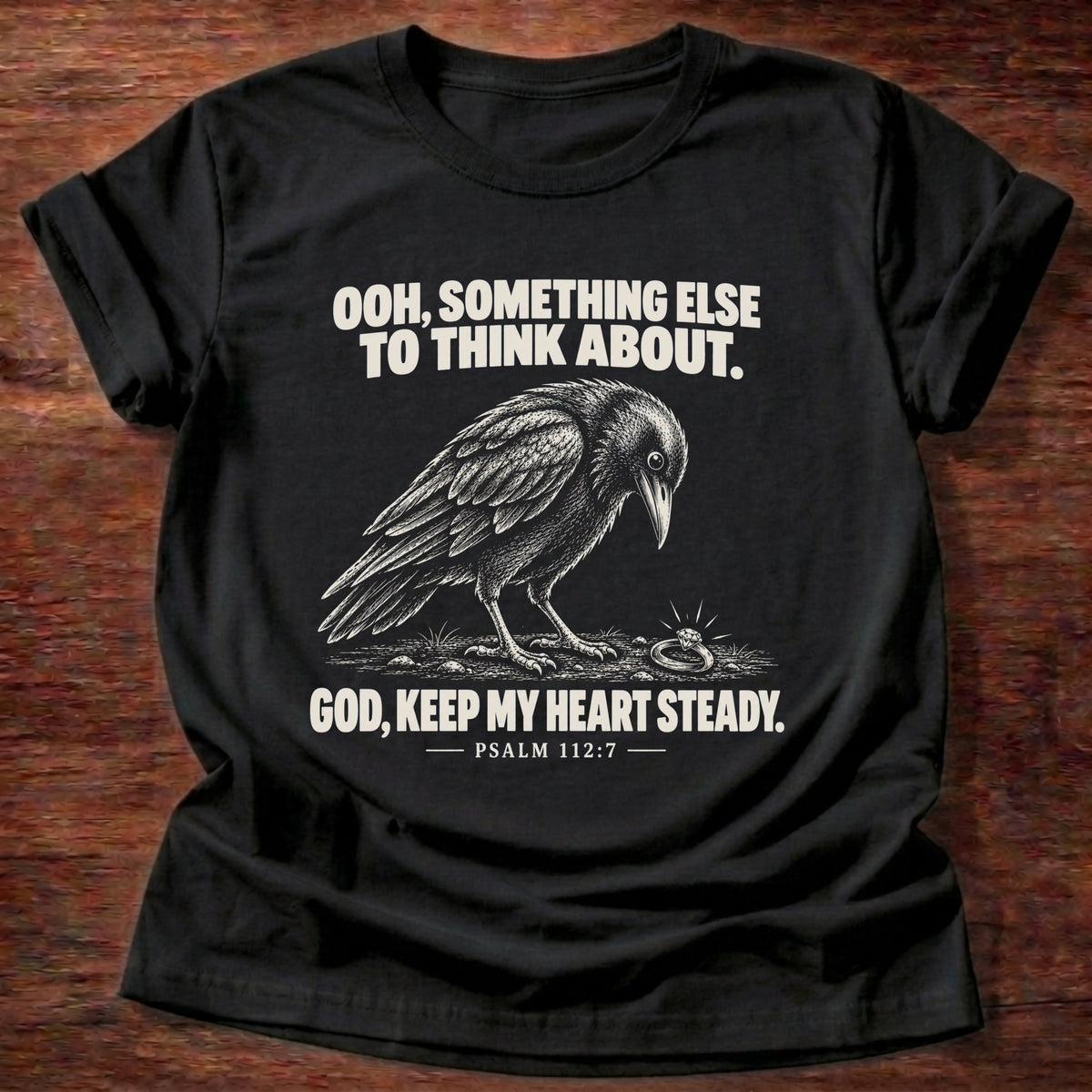 Black t-shirt printed with a crow investigating something shiny on the ground graphic and 'Ooh, something else to think about. God, keep my heart steady.' Psalm 112:7 faith based gift