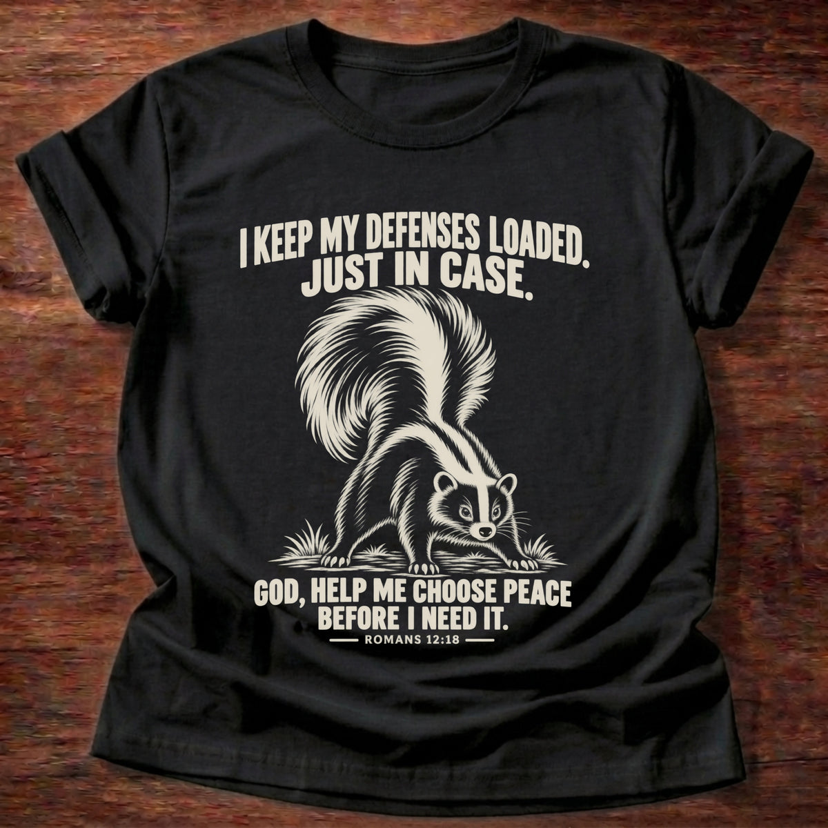 Black t-shirt printed with a skunk with its tail raised, ready to spray graphic and 'I keep my defenses loaded. Just in case. God, help me choose peace before I need it.' Romans 12:18 faith based gift