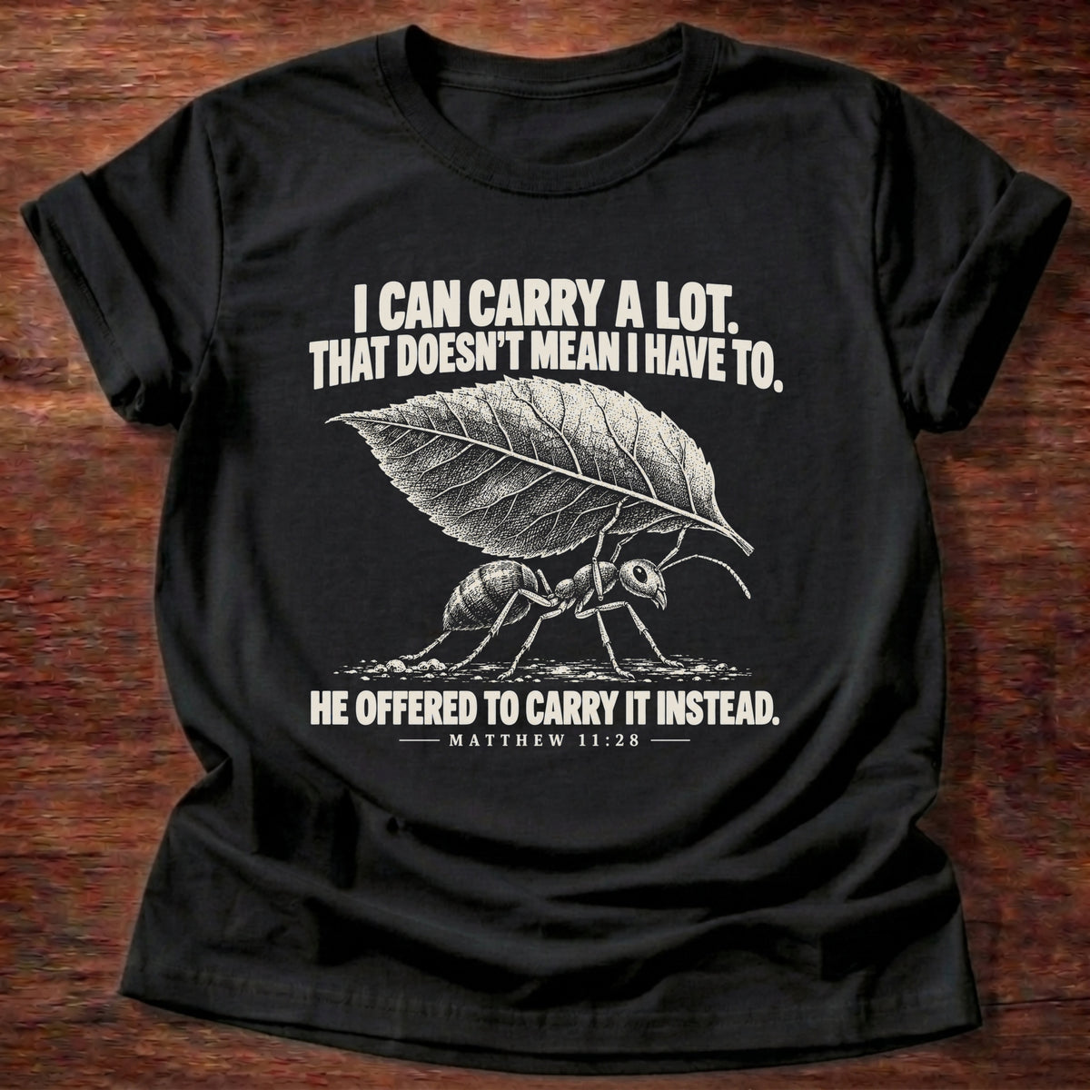 Black t-shirt printed with an ant hauling a leaf many times its size graphic and 'I can carry a lot. That doesn't mean I have to. He offered to carry it instead.' Matthew 11:28 faith based gift