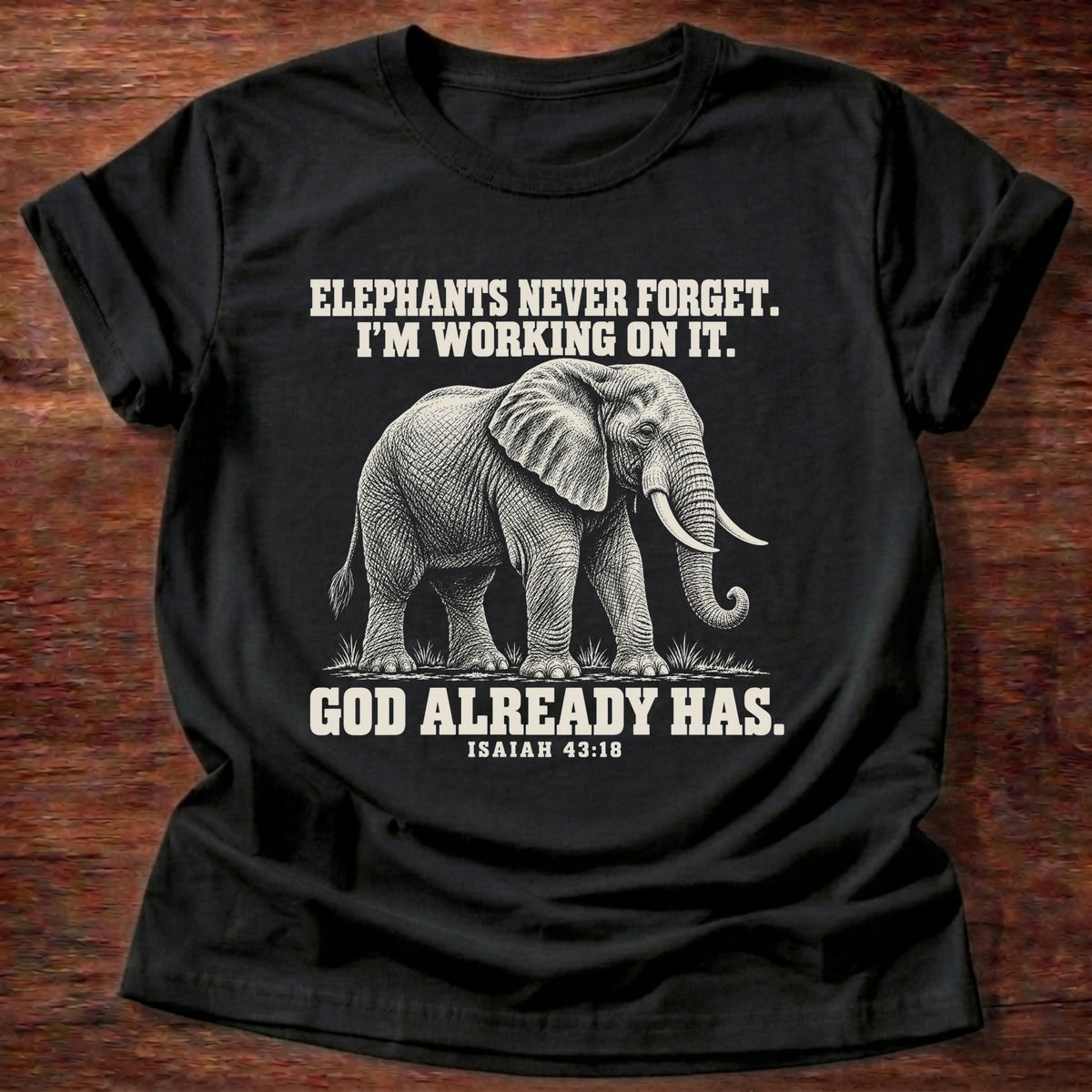 Black t-shirt printed with an elephant walking through tall grass graphic and 'Elephants never forget. I'm working on it. God already has.' Isaiah 43:18 faith based gift