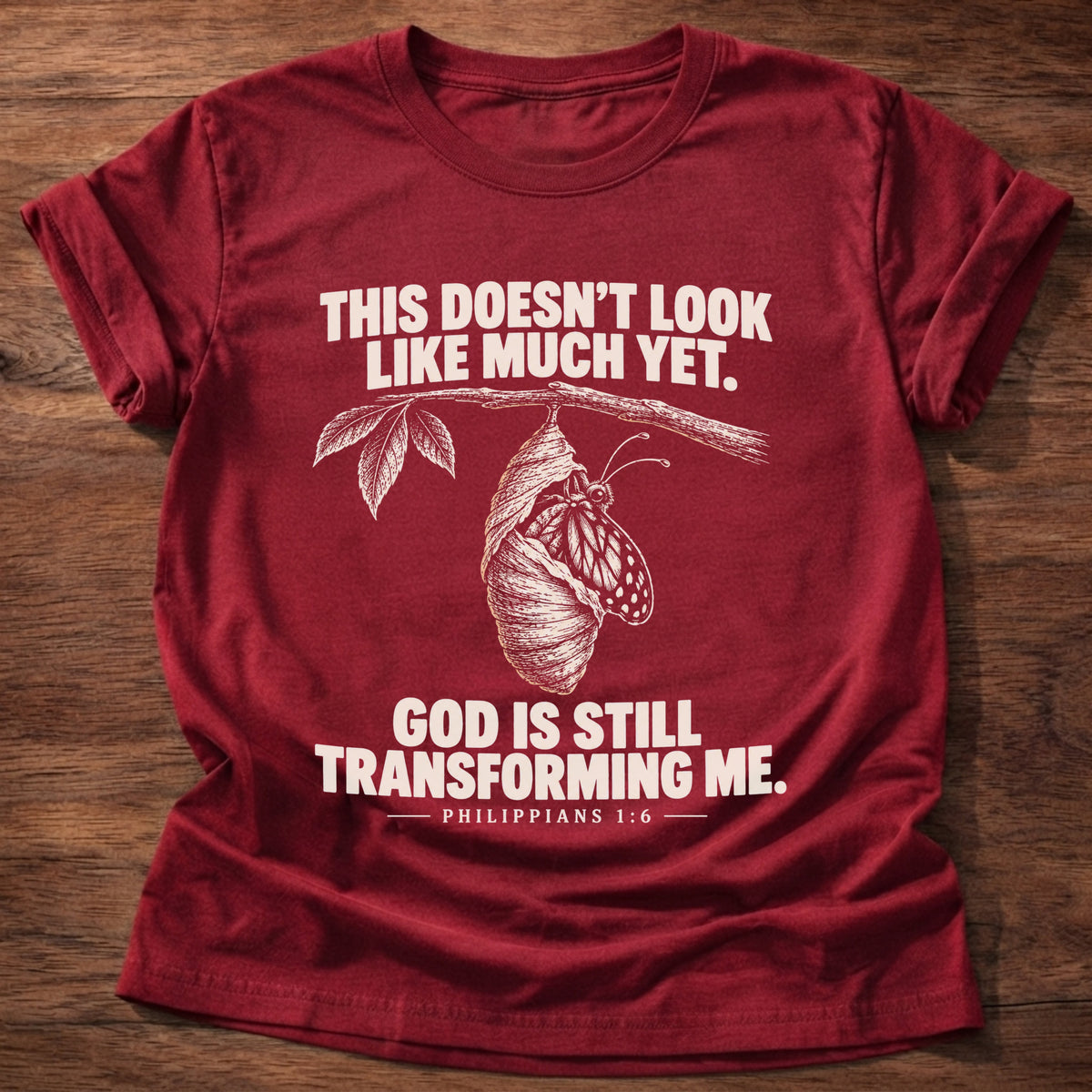 Cherry Red t-shirt printed with a chrysalis hanging from a branch graphic and 'This doesn't look like much yet. God is still transforming me.' Philippians 1:6 faith based gift