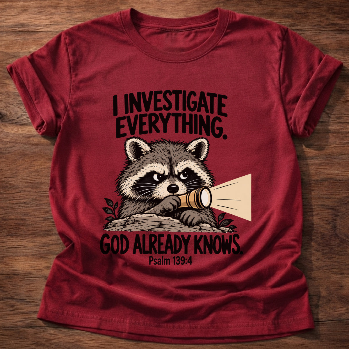 Cherry Red t-shirt printed with a raccoon holding a magnifying glass graphic and 'I investigate everything. God already knows.' Psalm 139:4 faith based gift
