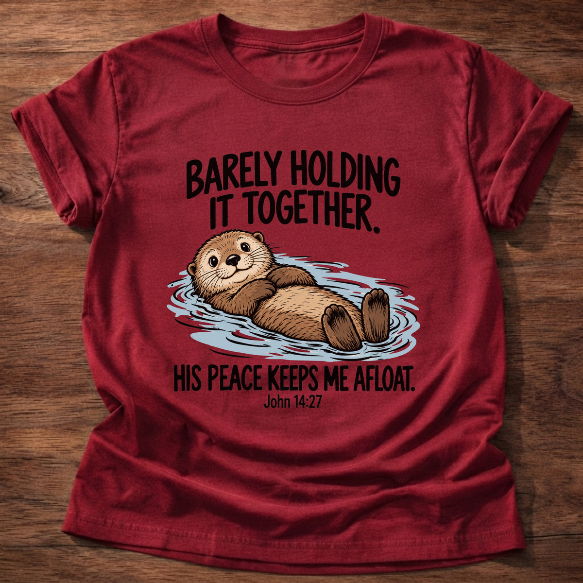 Cherry Red t-shirt printed with an otter floating on its back graphic and 'Barely holding it together. His peace keeps me afloat.' John 14:27 faith based gift