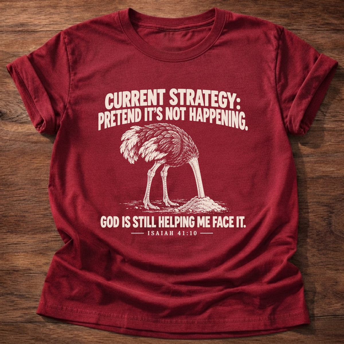 Cherry Red t-shirt printed with an ostrich with its head buried in the sand graphic and 'Current strategy: pretend it's not happening. God is still helping me face it.' Isaiah 41:10 faith based gift