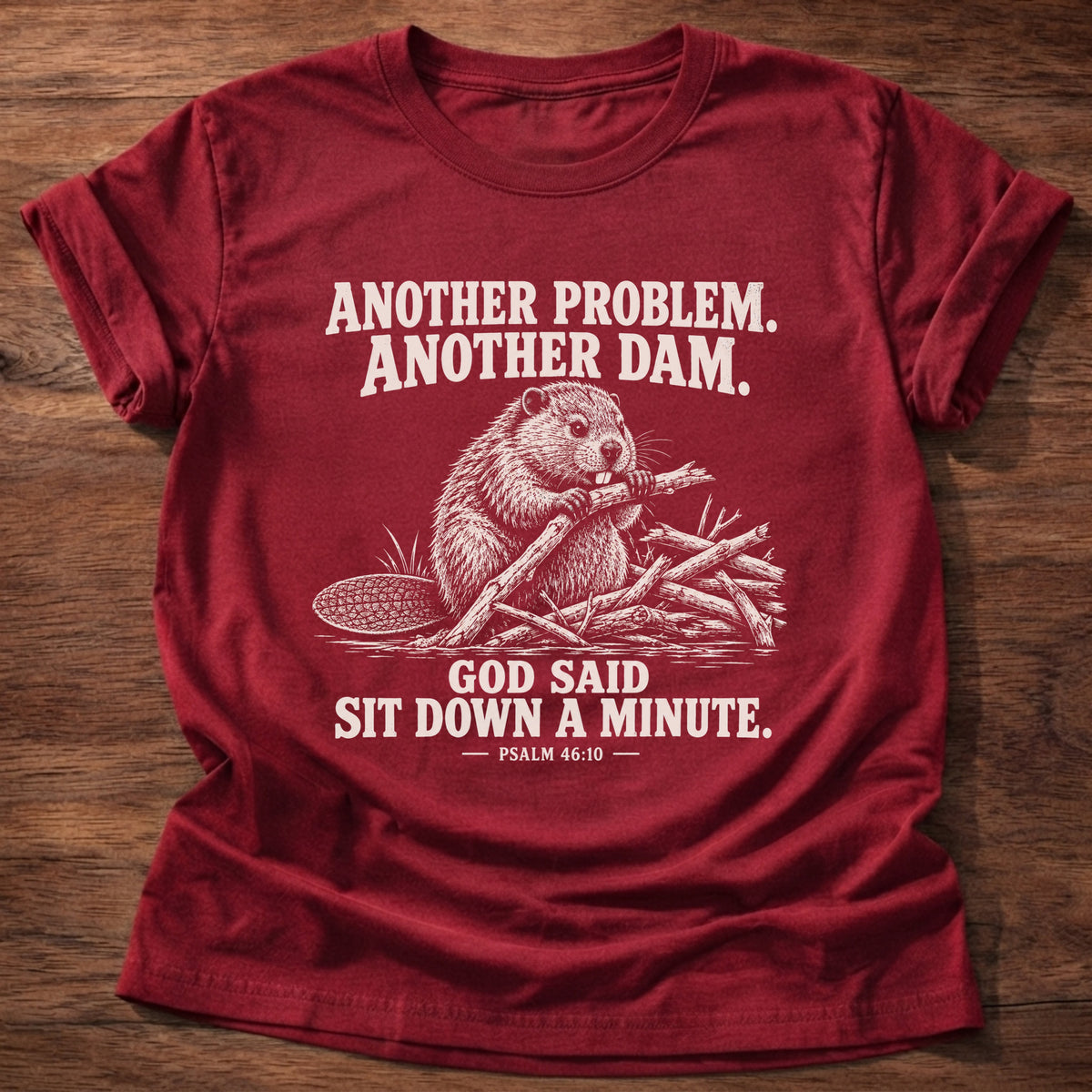 Cherry Red t-shirt printed with a beaver mid-build with another branch graphic and 'Another problem. Another dam. God said sit down a minute.' Psalm 46:10 faith based gift