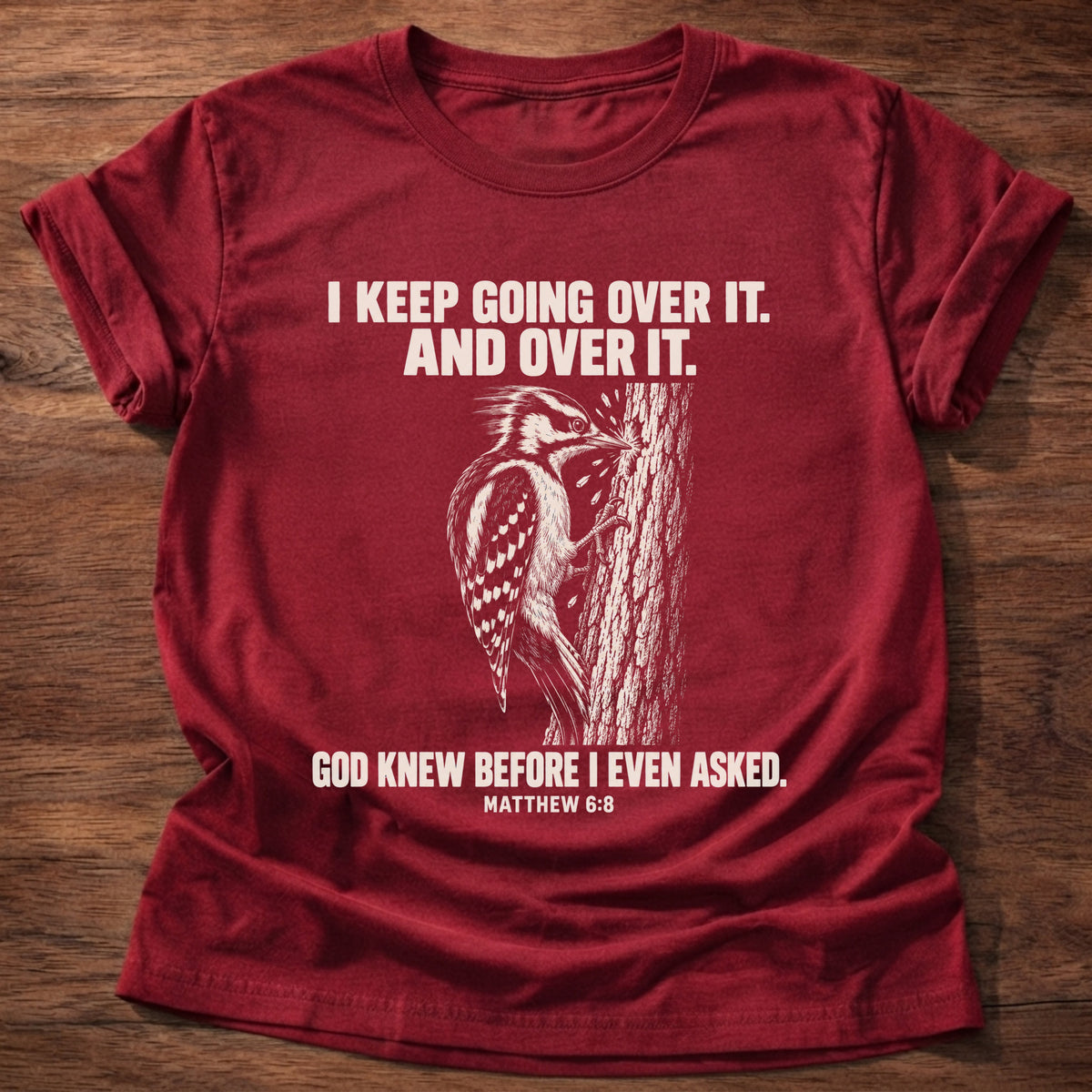 Cherry Red t-shirt printed with a woodpecker drumming the same spot on a tree graphic and 'I keep going over it. And over it. God knew before I even asked.' Matthew 6:8 faith based gift