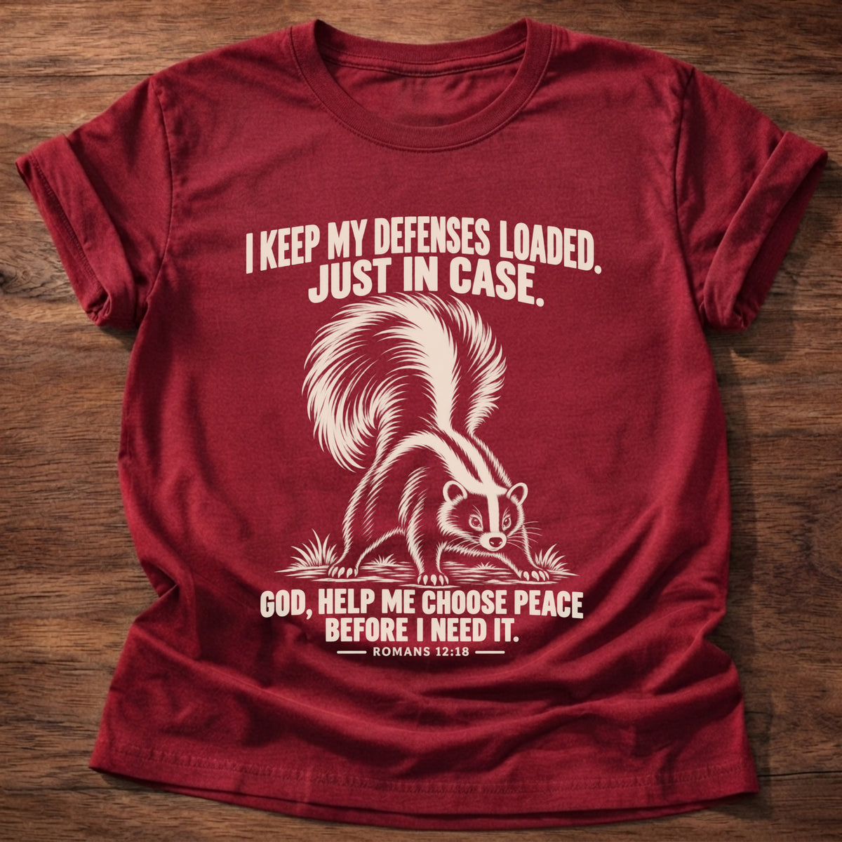 Cherry Red t-shirt printed with a skunk with its tail raised, ready to spray graphic and 'I keep my defenses loaded. Just in case. God, help me choose peace before I need it.' Romans 12:18 faith based gift