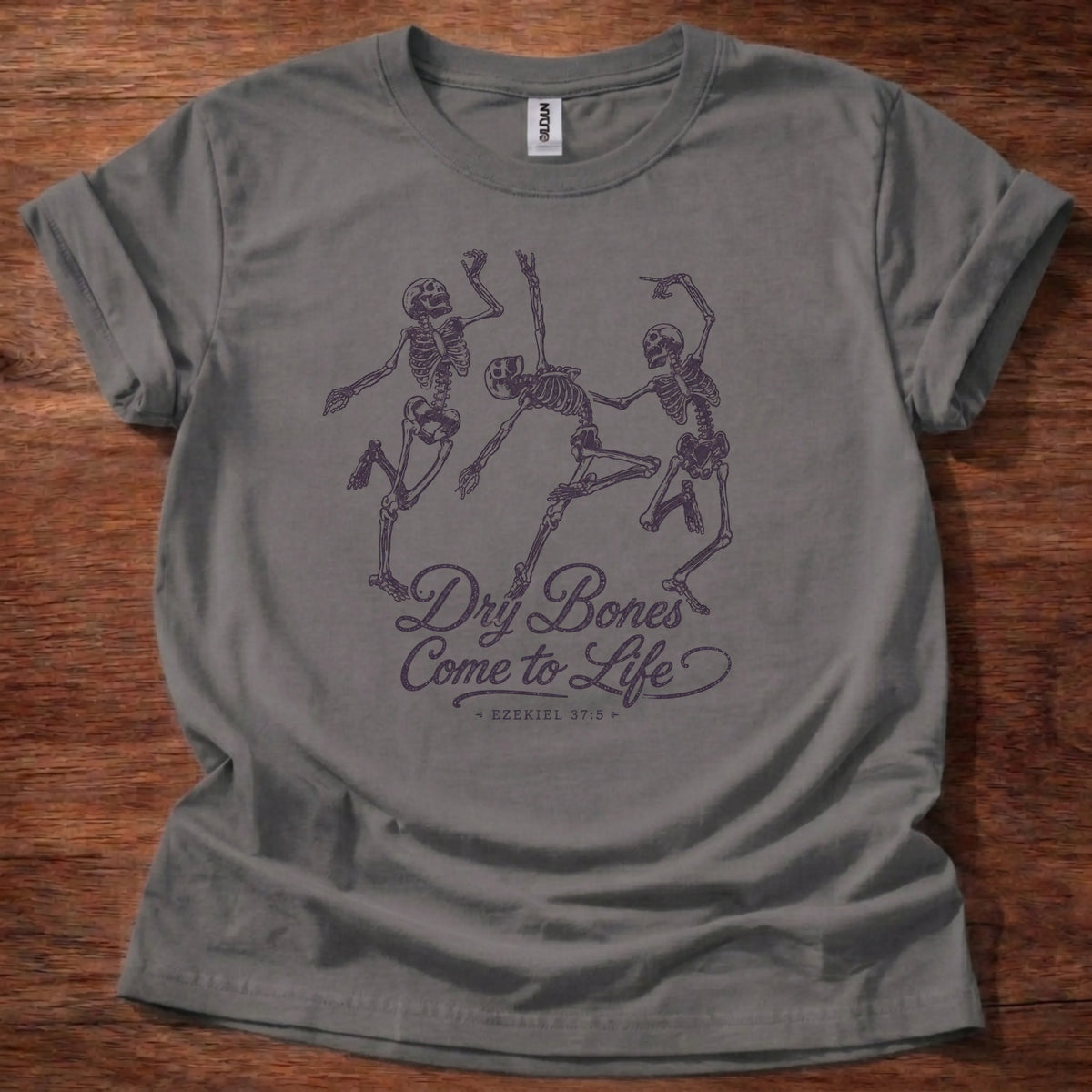 Charcoal t-shirt with three skeletons dancing in vintage tattoo-flash linework above cursive text reading Dry Bones Come to Life, Ezekiel 37:5