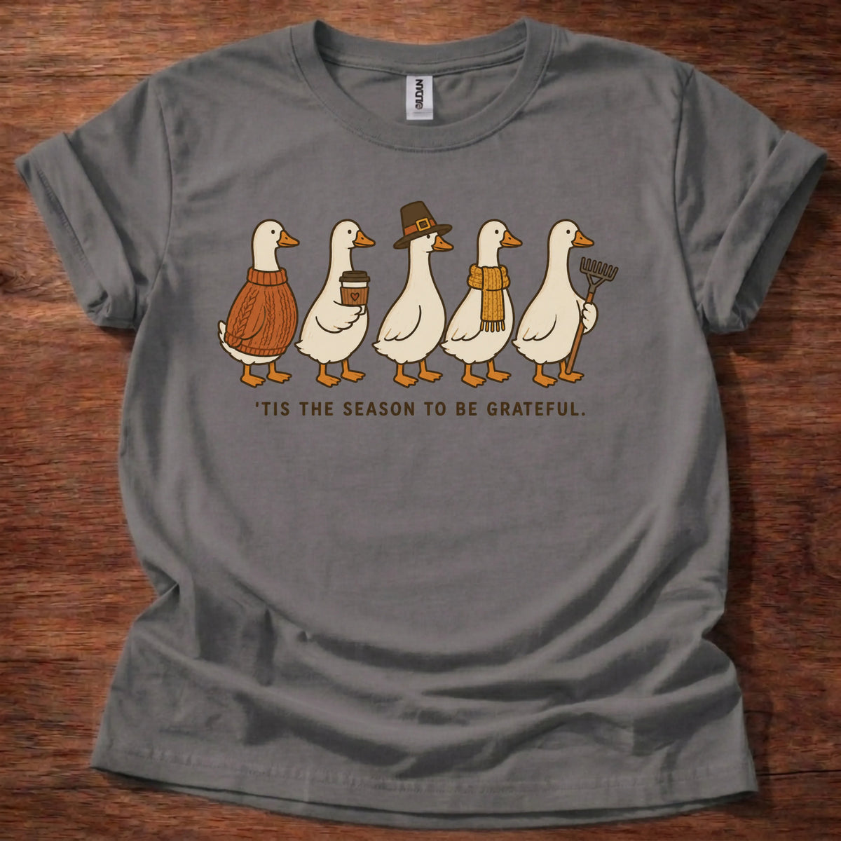 Charcoal t-shirt with five folk-art geese wearing sweaters, a pilgrim hat, and a scarf above text reading 'Tis the Season to Be Grateful