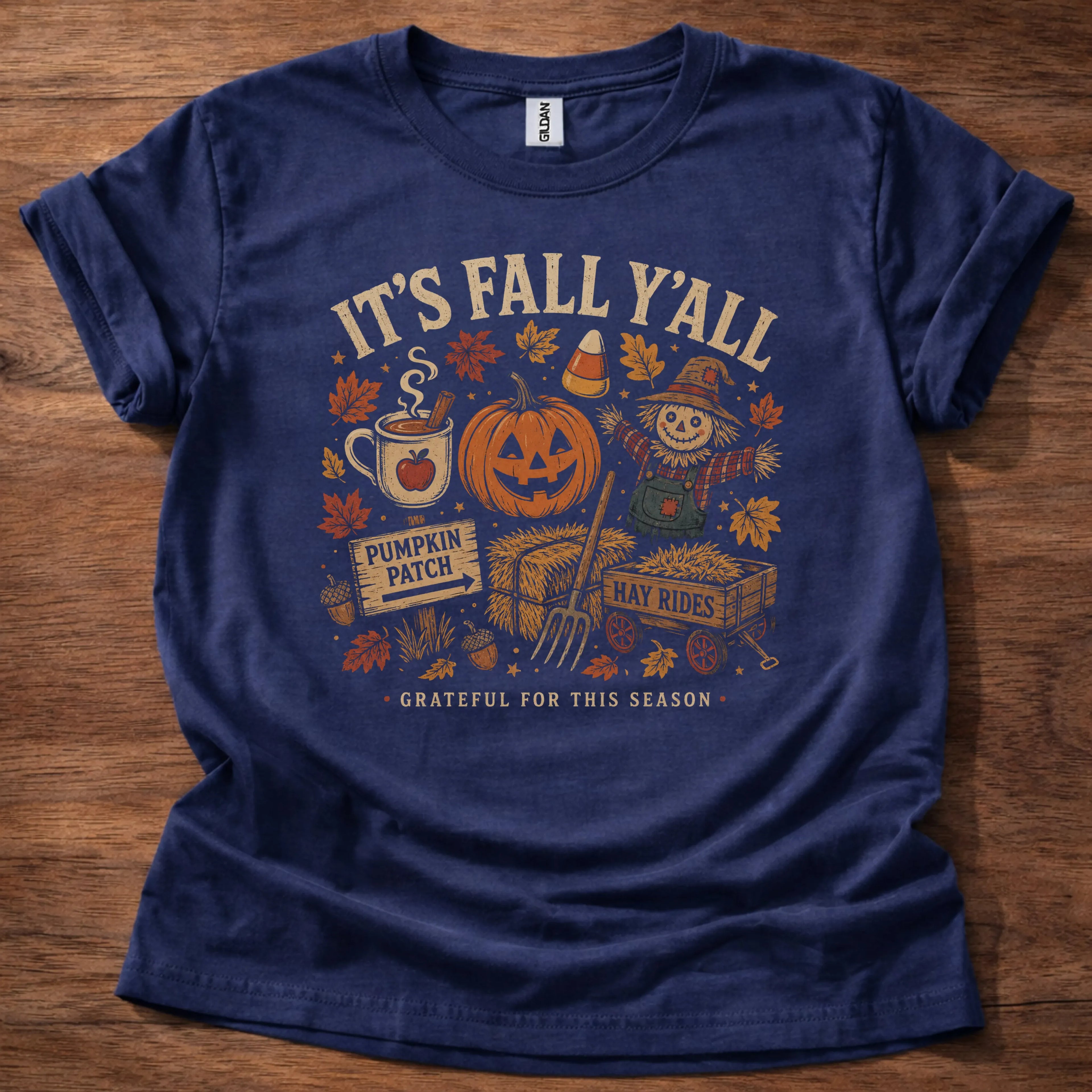 Navy t-shirt with a bold flash-art cluster of fall icons including a pumpkin, cider mug, scarecrow, and hay rides sign around text reading It's Fall Y'all, Grateful For This Season