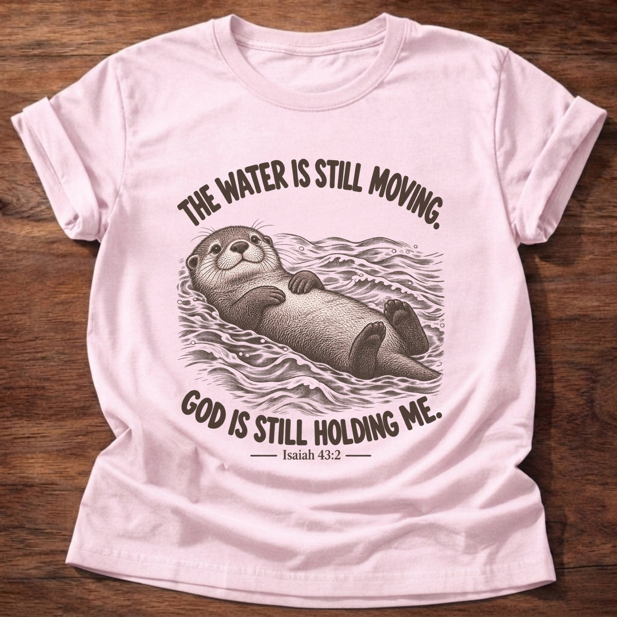 Light Pink t-shirt printed with an otter floating on its back in moving water graphic and “The water is still moving. God is still holding me.” Isaiah 43:2 faith based gift