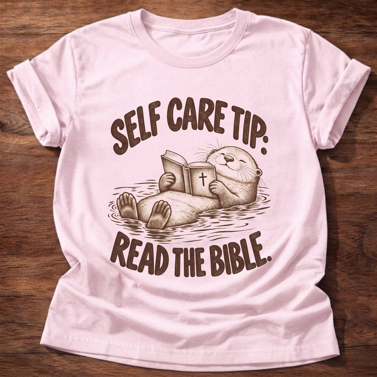 Light Pink t-shirt printed with a sea otter floating on its back reading a Bible graphic and “Self care tip: Read the Bible.” faith based gift