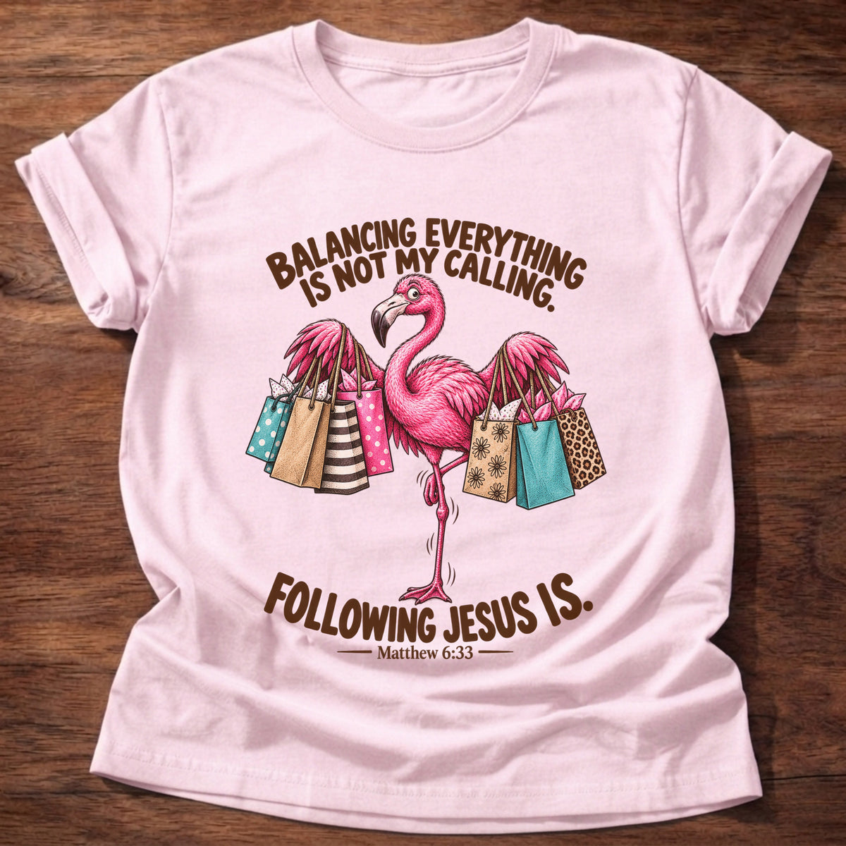 Light Pink t-shirt printed with a pink flamingo balancing on one leg loaded with colorful shopping bags graphic and “Balancing everything is not my calling. Following Jesus is.” Matthew 6:33 faith based gift