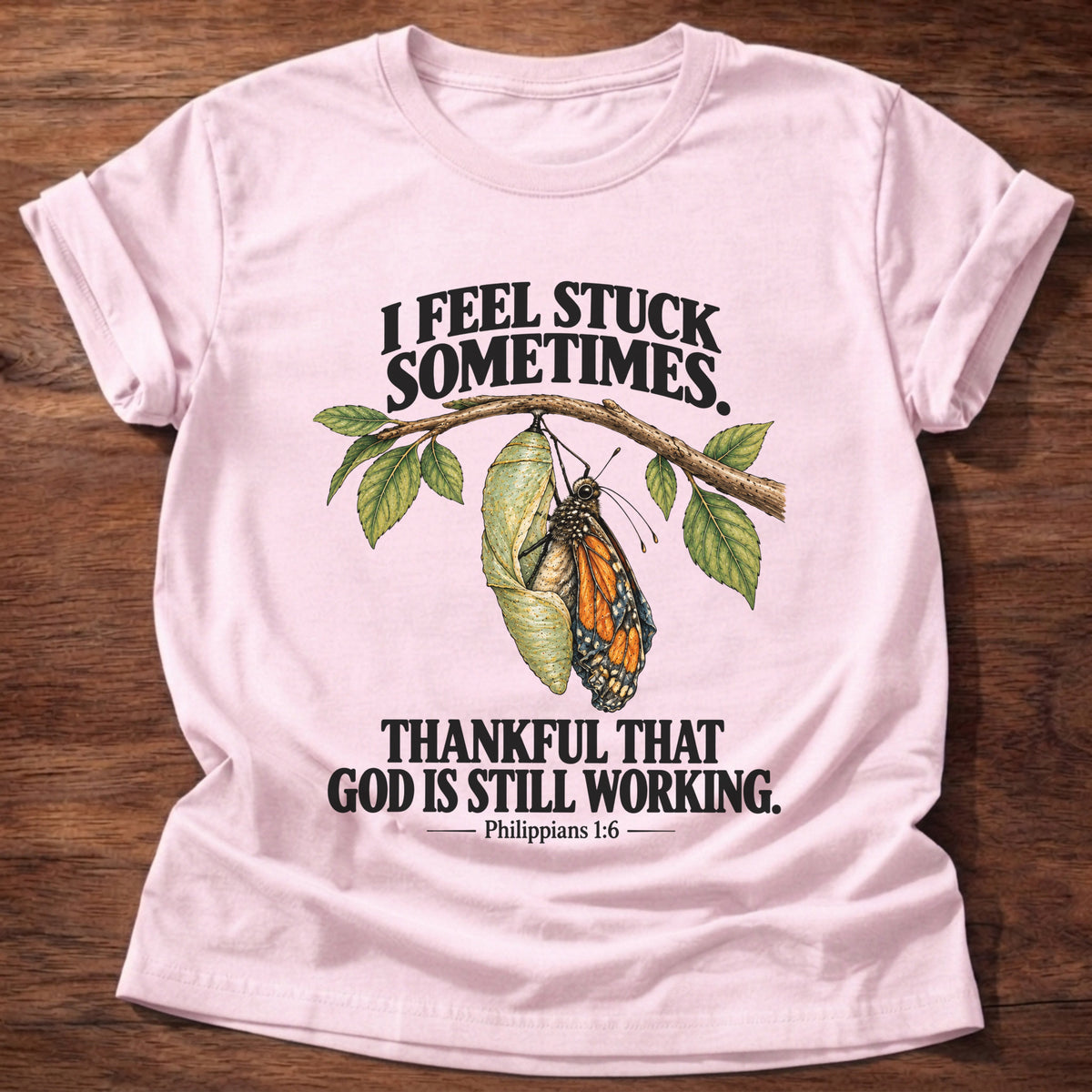 Light Pink t-shirt printed with a monarch butterfly emerging from its chrysalis on a leafy branch graphic and “I feel stuck sometimes. Thankful that God is still working.” Philippians 1:6 faith based gift
