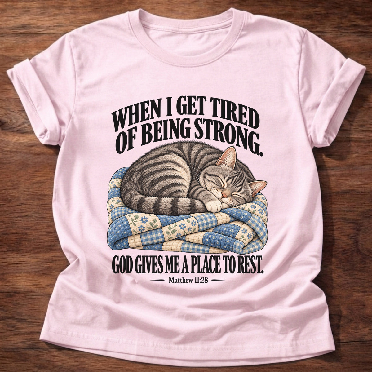 Light Pink t-shirt printed with a tabby cat curled up asleep on a folded patchwork quilt graphic and “When I get tired of being strong, God gives me a place to rest.” Matthew 11:28 faith based gift