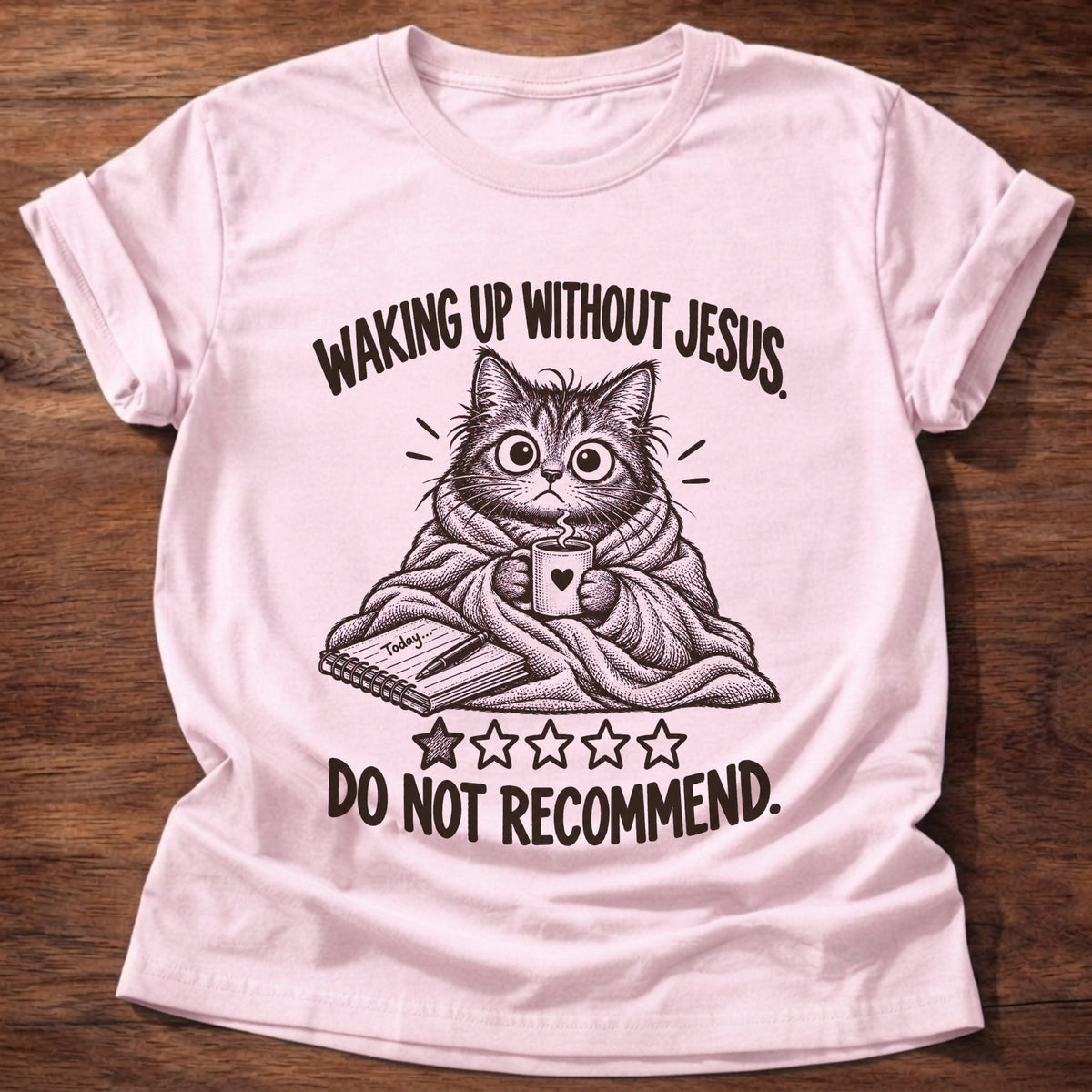 Light Pink t-shirt printed with a frazzled wide-eyed cat wrapped in a blanket holding a coffee mug above a one-star rating graphic and “Waking up without Jesus. Do not recommend.” faith based gift