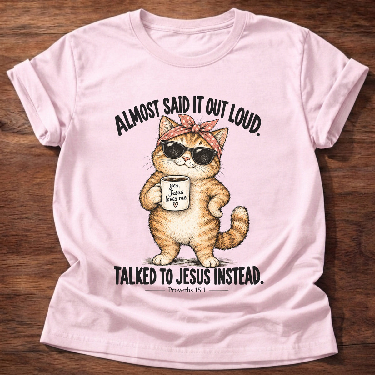 Light Pink t-shirt printed with an orange tabby cat in sunglasses and a polka-dot bandana holding a 'yes, Jesus loves me' mug graphic and “Almost said it out loud. Talked to Jesus instead.” Proverbs 15:1 faith based gift