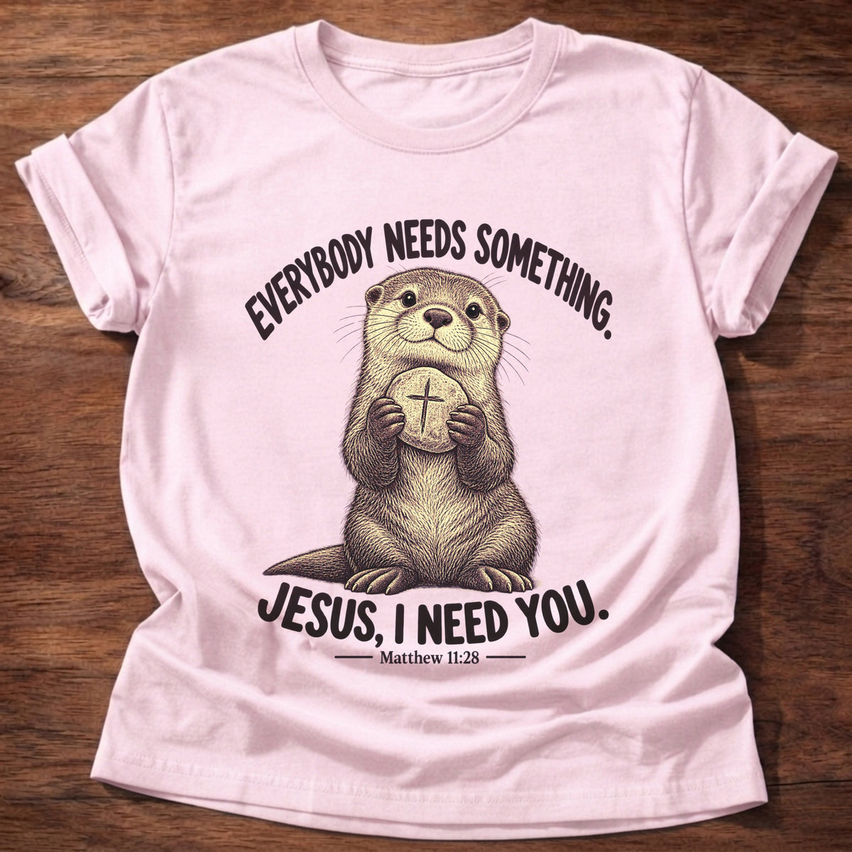 Light Pink t-shirt printed with an otter hugging a round stone etched with a cross graphic and “Everybody needs something. Jesus, I need you.” Matthew 11:28 faith based gift