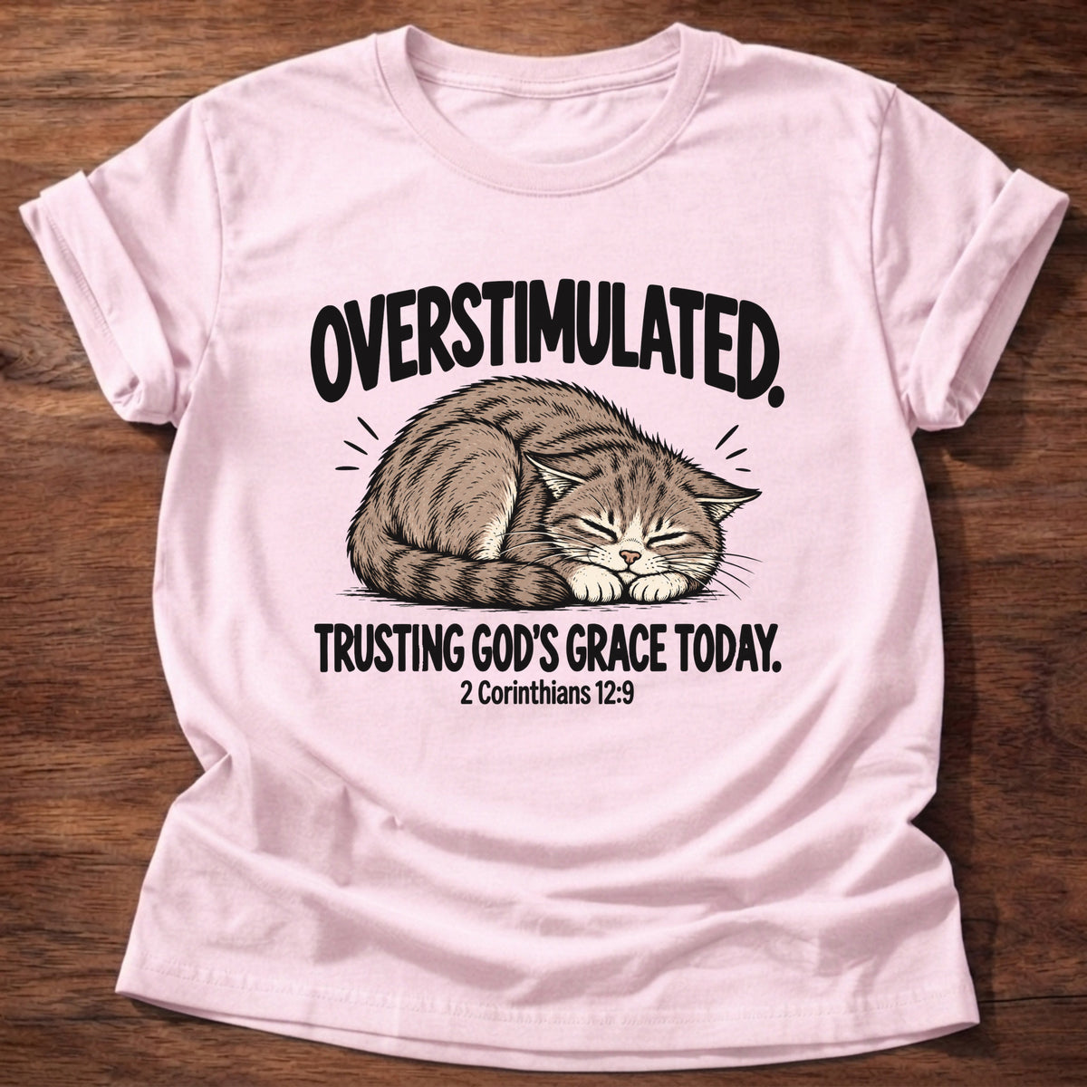 Light Pink t-shirt printed with a cat curled asleep graphic and 'Overstimulated. Trusting God's grace today.' 2 Corinthians 12:9 faith based gift