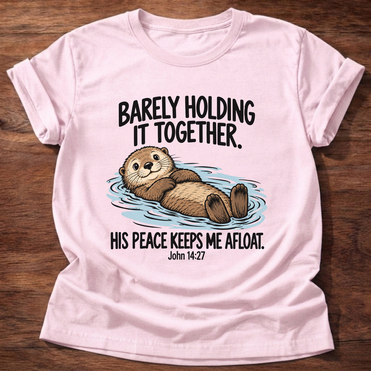 Light Pink t-shirt printed with an otter floating on its back graphic and 'Barely holding it together. His peace keeps me afloat.' John 14:27 faith based gift