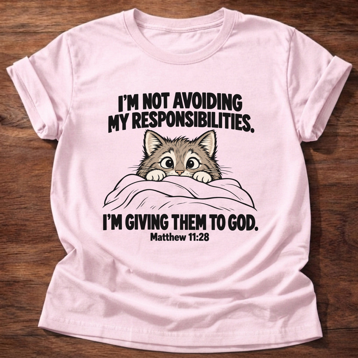 Light Pink t-shirt printed with a cat peeking over a blanket graphic and 'I'm not avoiding my responsibilities. I'm giving them to God.' Matthew 11:28 faith based gift