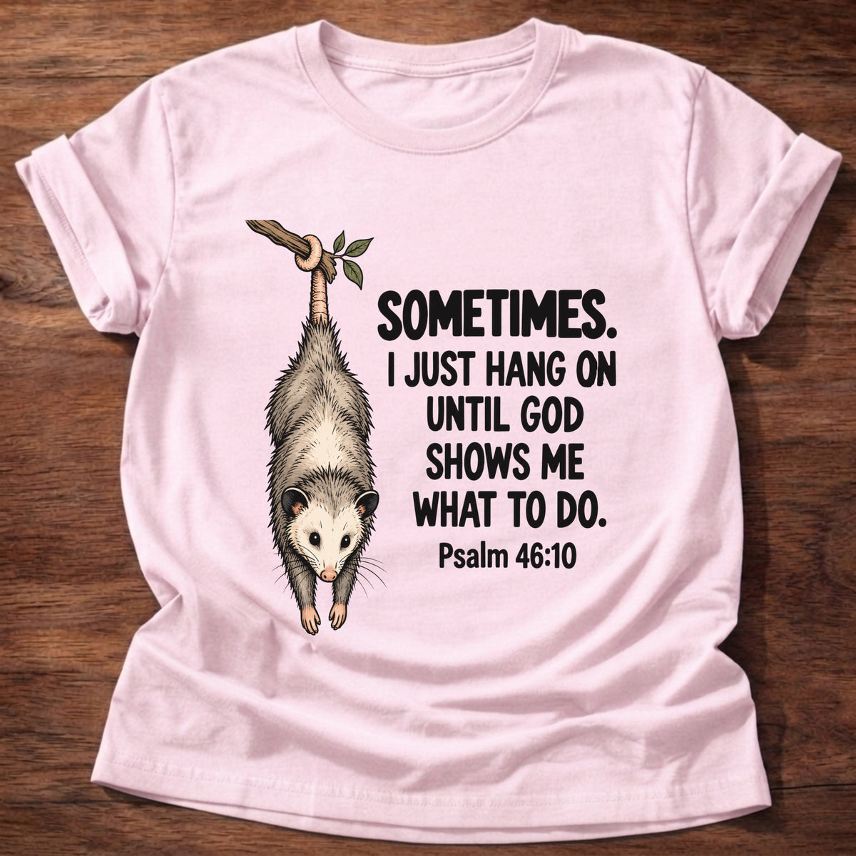 Light Pink t-shirt printed with a possum hanging by its tail graphic and 'Sometimes. I just hang on until God shows me what to do.' Psalm 46:10 faith based gift