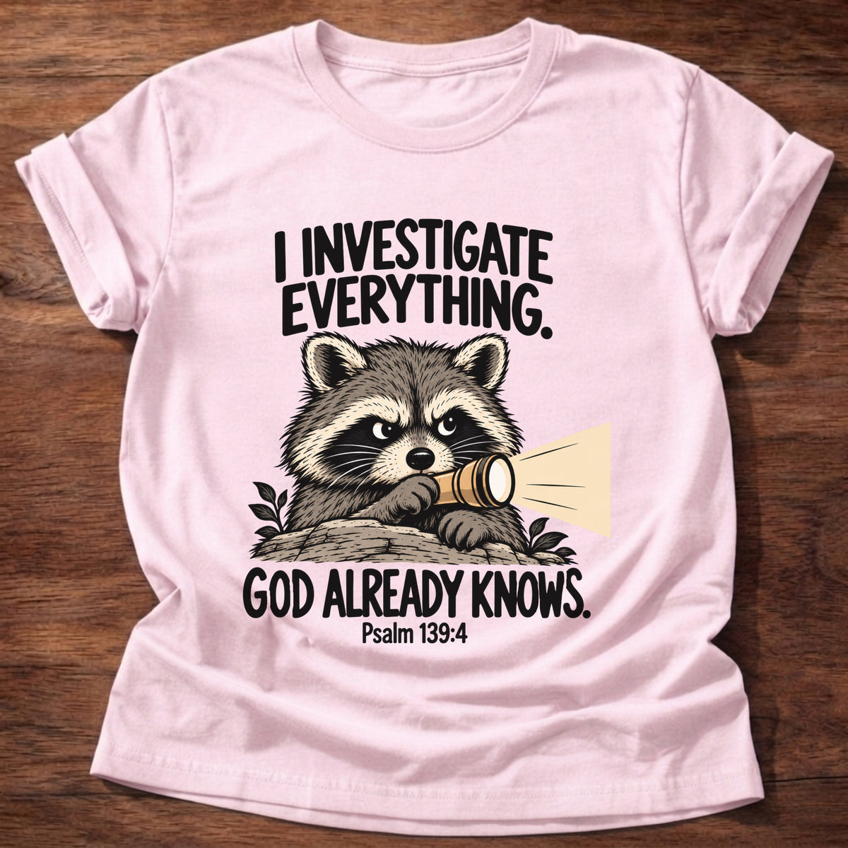 Light Pink t-shirt printed with a raccoon holding a magnifying glass graphic and 'I investigate everything. God already knows.' Psalm 139:4 faith based gift