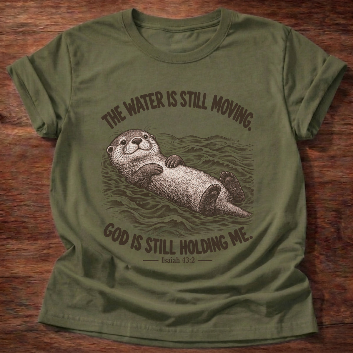 Military Green t-shirt printed with an otter floating on its back in moving water graphic and “The water is still moving. God is still holding me.” Isaiah 43:2 faith based gift