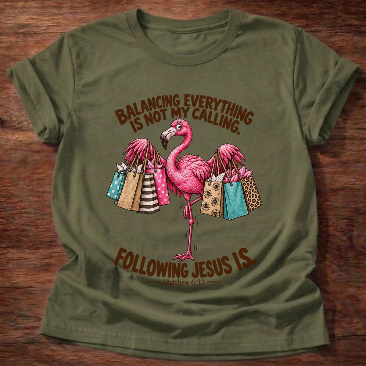 Military Green t-shirt printed with a pink flamingo balancing on one leg loaded with colorful shopping bags graphic and “Balancing everything is not my calling. Following Jesus is.” Matthew 6:33 faith based gift