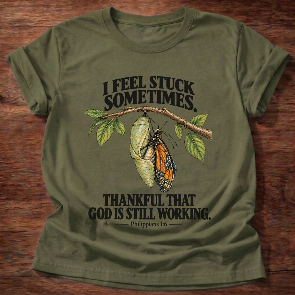 Military Green t-shirt printed with a monarch butterfly emerging from its chrysalis on a leafy branch graphic and “I feel stuck sometimes. Thankful that God is still working.” Philippians 1:6 faith based gift
