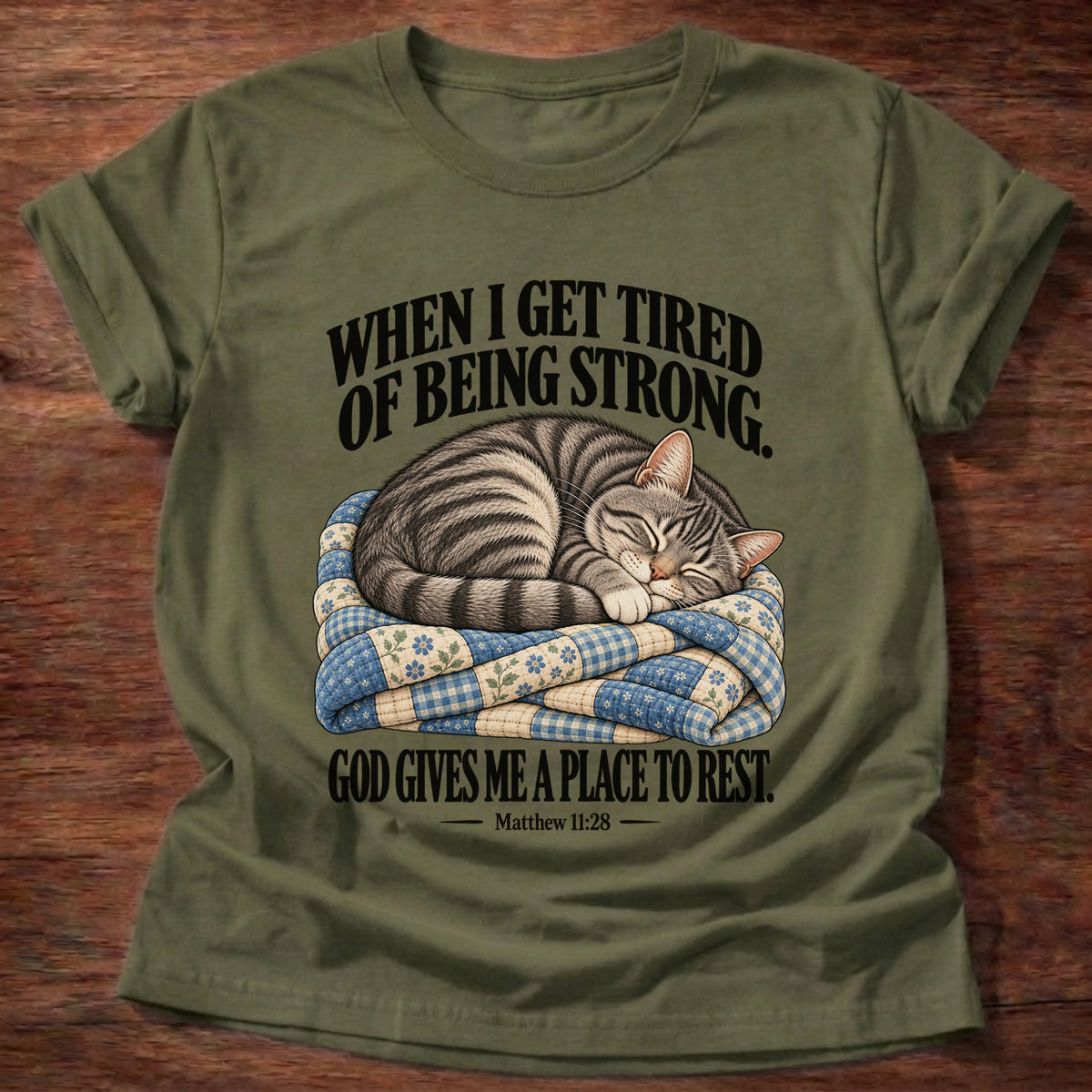 Military Green t-shirt printed with a tabby cat curled up asleep on a folded patchwork quilt graphic and “When I get tired of being strong, God gives me a place to rest.” Matthew 11:28 faith based gift