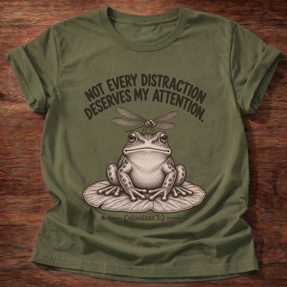 Military Green t-shirt printed with a frog sitting on a lily pad with a dragonfly perched on its head graphic and “Not every distraction deserves my attention.” Colossians 3:2 faith based gift