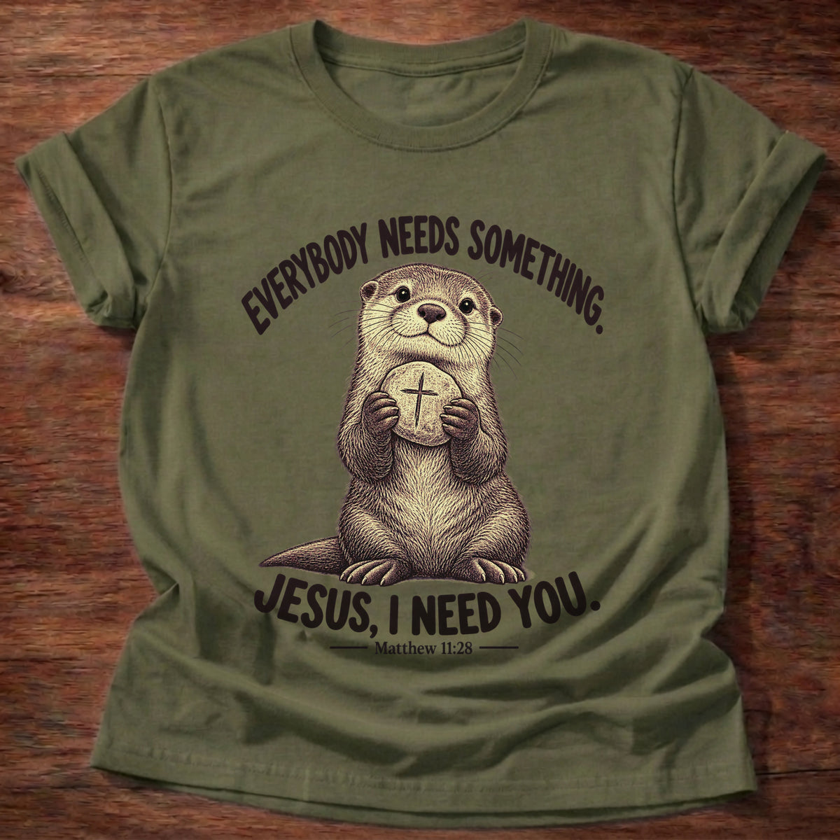 Military Green t-shirt printed with an otter hugging a round stone etched with a cross graphic and “Everybody needs something. Jesus, I need you.” Matthew 11:28 faith based gift