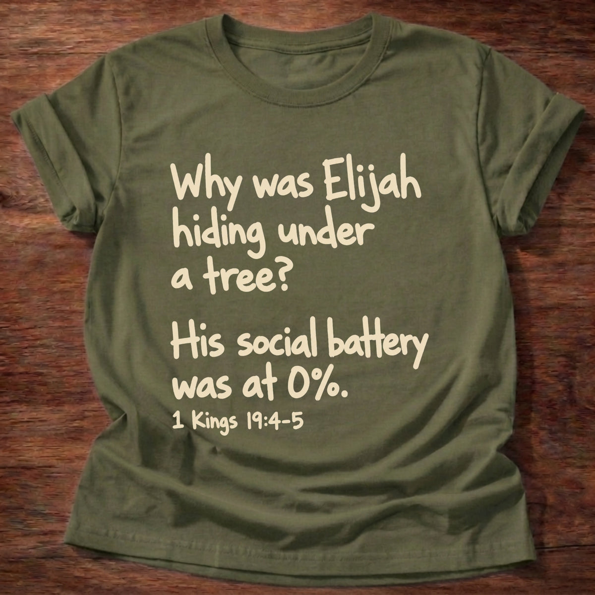 Military Green t-shirt printed with a simple hand-lettered Bible joke graphic and “Why was Elijah hiding under a tree? His social battery was at 0%.” 1 Kings 19:4-5 Christian shirt