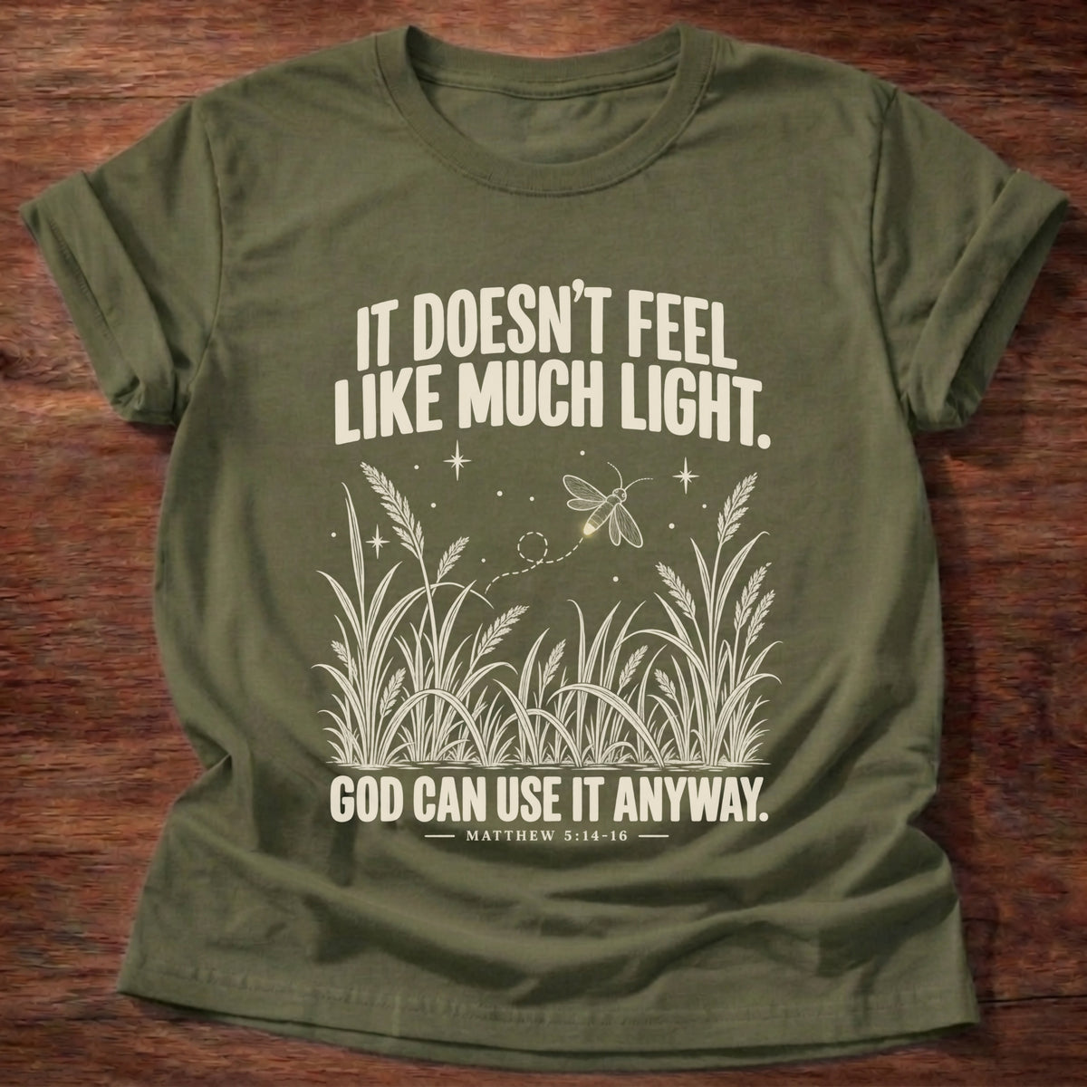 Military Green t-shirt printed with a firefly glowing in a dark field of grass graphic and 'It doesn't feel like much light. God can use it anyway.' Matthew 5:14-16 faith based gift