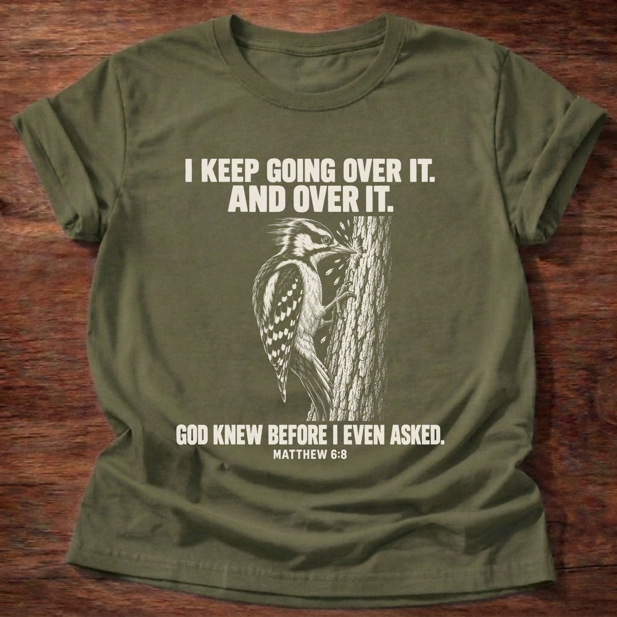 Military Green t-shirt printed with a woodpecker drumming the same spot on a tree graphic and 'I keep going over it. And over it. God knew before I even asked.' Matthew 6:8 faith based gift
