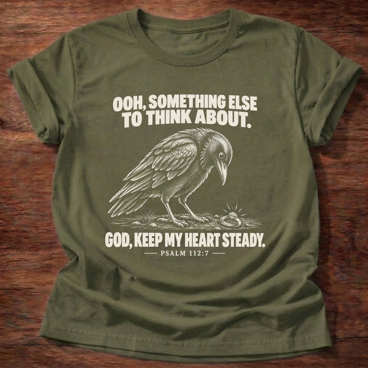 Military Green t-shirt printed with a crow investigating something shiny on the ground graphic and 'Ooh, something else to think about. God, keep my heart steady.' Psalm 112:7 faith based gift